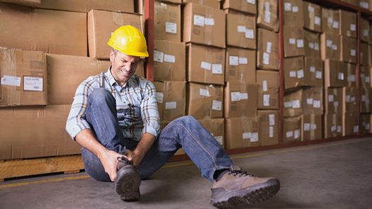 A man in a hard hat sits near a wall of boxes clutching his ankle in pain
